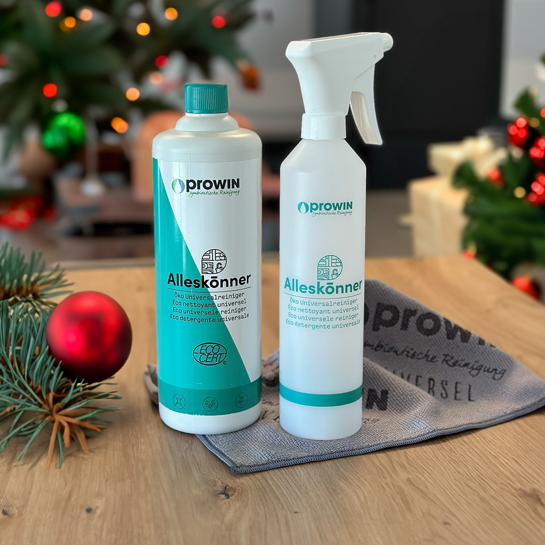 Deux bouteilles de nettoyant universel Prowin "Alleskönner", nettoyant à tout faire, sont posées sur une table en bois, accompagnées d’un chiffon gris avec le logo de la marque. L’arrière-plan festif révèle un sapin de Noël décoré et des guirlandes lumineuses, créant une ambiance chaleureuse et conviviale. Une branche de sapin ornée d’une boule rouge ajoute une touche festive à la scène.