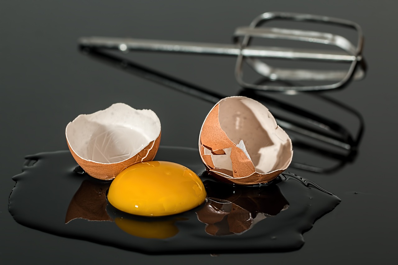 Un œuf cassé avec son jaune éclaté repose sur une surface noire brillante, entouré de coquilles brisées. À l'arrière-plan, des fouets métalliques d’un batteur soulignent l’ambiance de préparation culinaire.