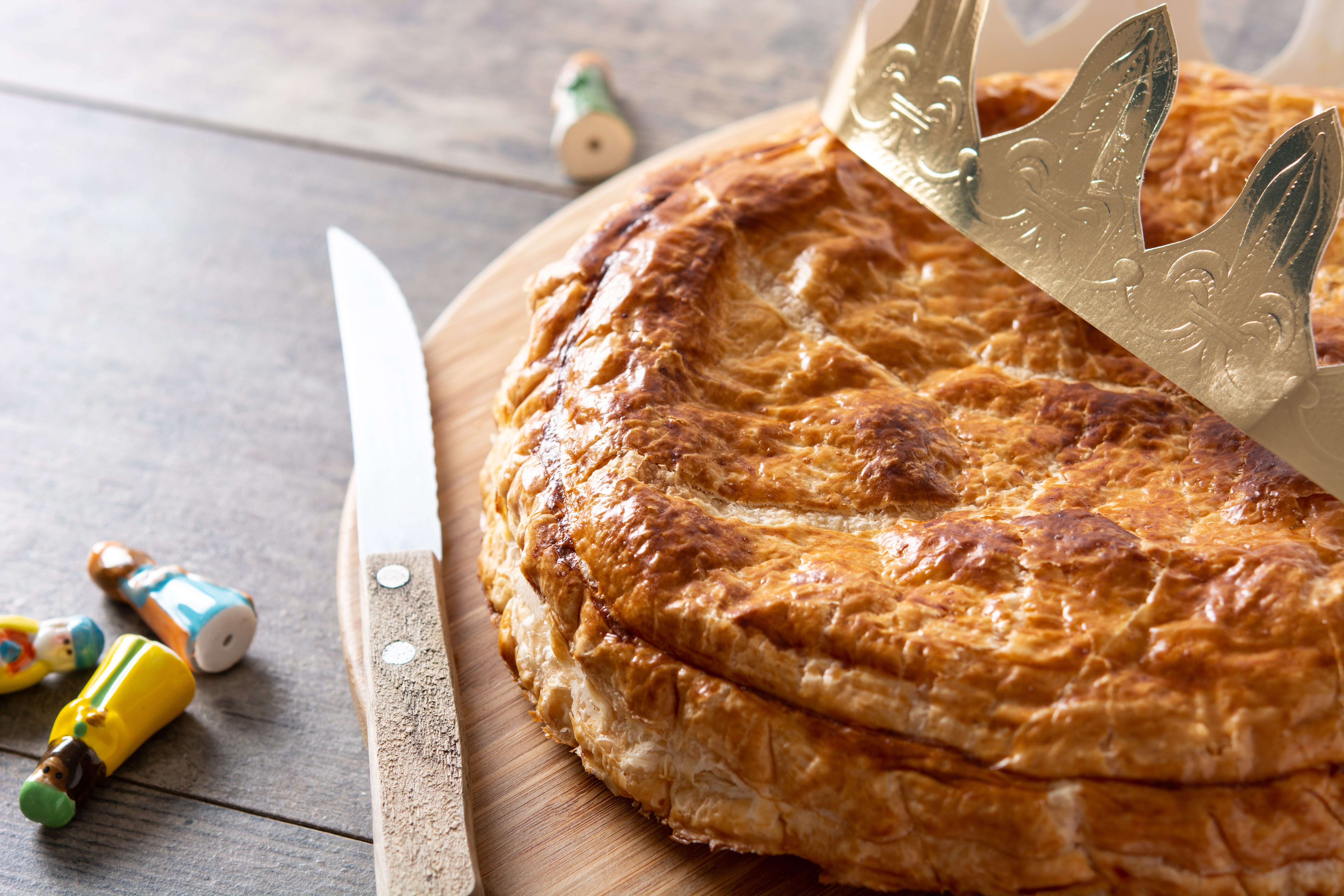 Un Pithiviers doré repose sur une planche en bois, couronné d'une élégante couronne dorée. À côté, un couteau et quelques fèves colorées ajoutent une touche festive, évoquant la tradition de l’Épiphanie.