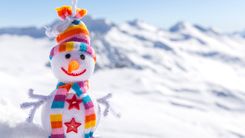 Un bonhomme de neige coloré avec une écharpe et un bonnet tricotés, posé sur une neige immaculée avec un paysage de montagnes enneigées en arrière-plan.