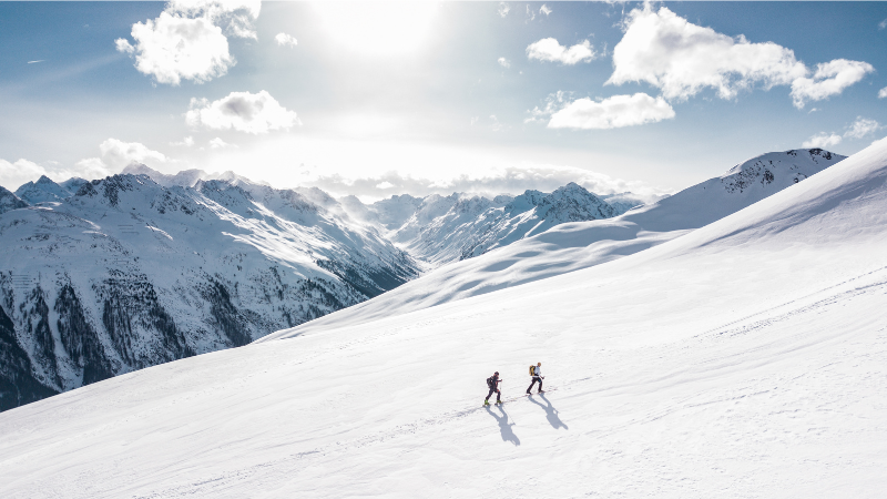 Deux randonneurs progressent sur une pente enneigée en haute montagne, sous un ciel bleu et ensoleillé, avec une vue panoramique sur des sommets enneigés à l'horizon.