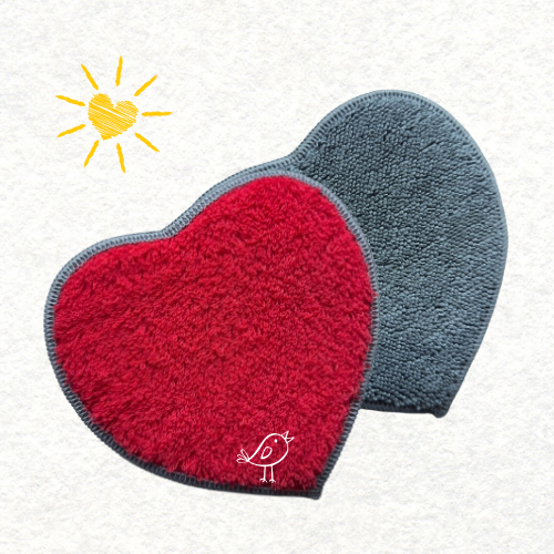 Éponges en forme de cœur, l’une rouge et l’autre gris anthracite, posées côte à côte sur fond clair avec un petit oiseau dessiné et un soleil en forme de cœur jaune dans le coin supérieur gauche. Accessoire de ménage doux et écologique, idéal pour nettoyer avec amour.